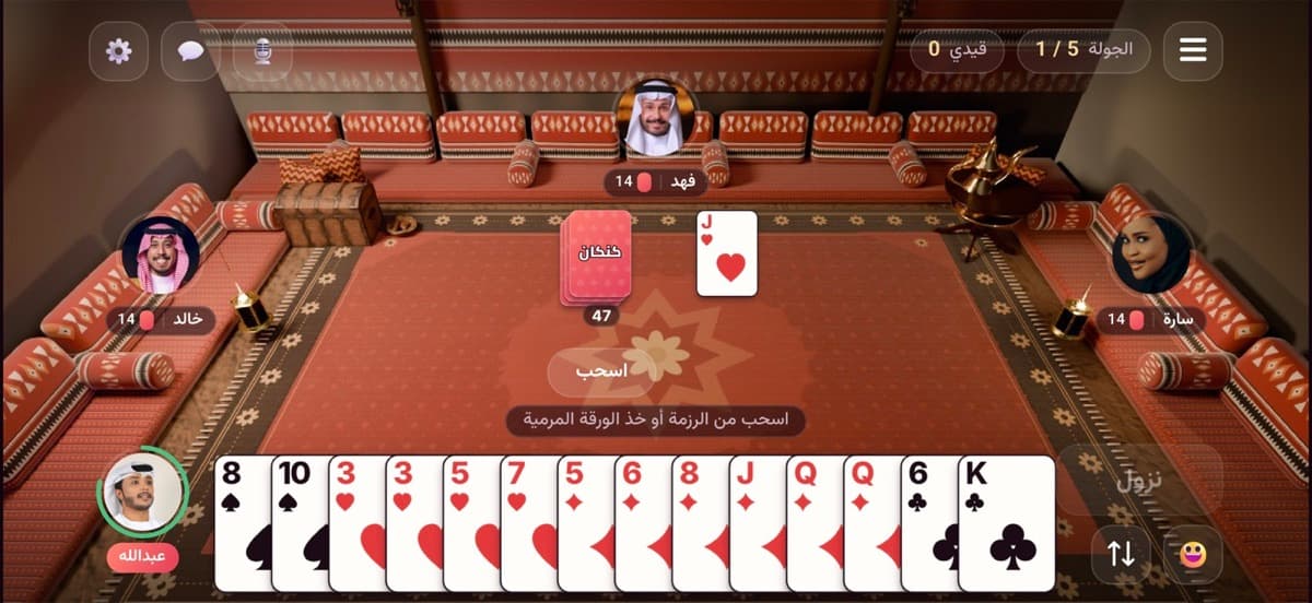 طاولةُ كنكان داخل اللعبة: أربعةُ لاعبين حول مجلسٍ خليجيّ، الرزمة والورقةُ المرميّة في الوسط ويدُ اللاعب أسفل الشاشة.
